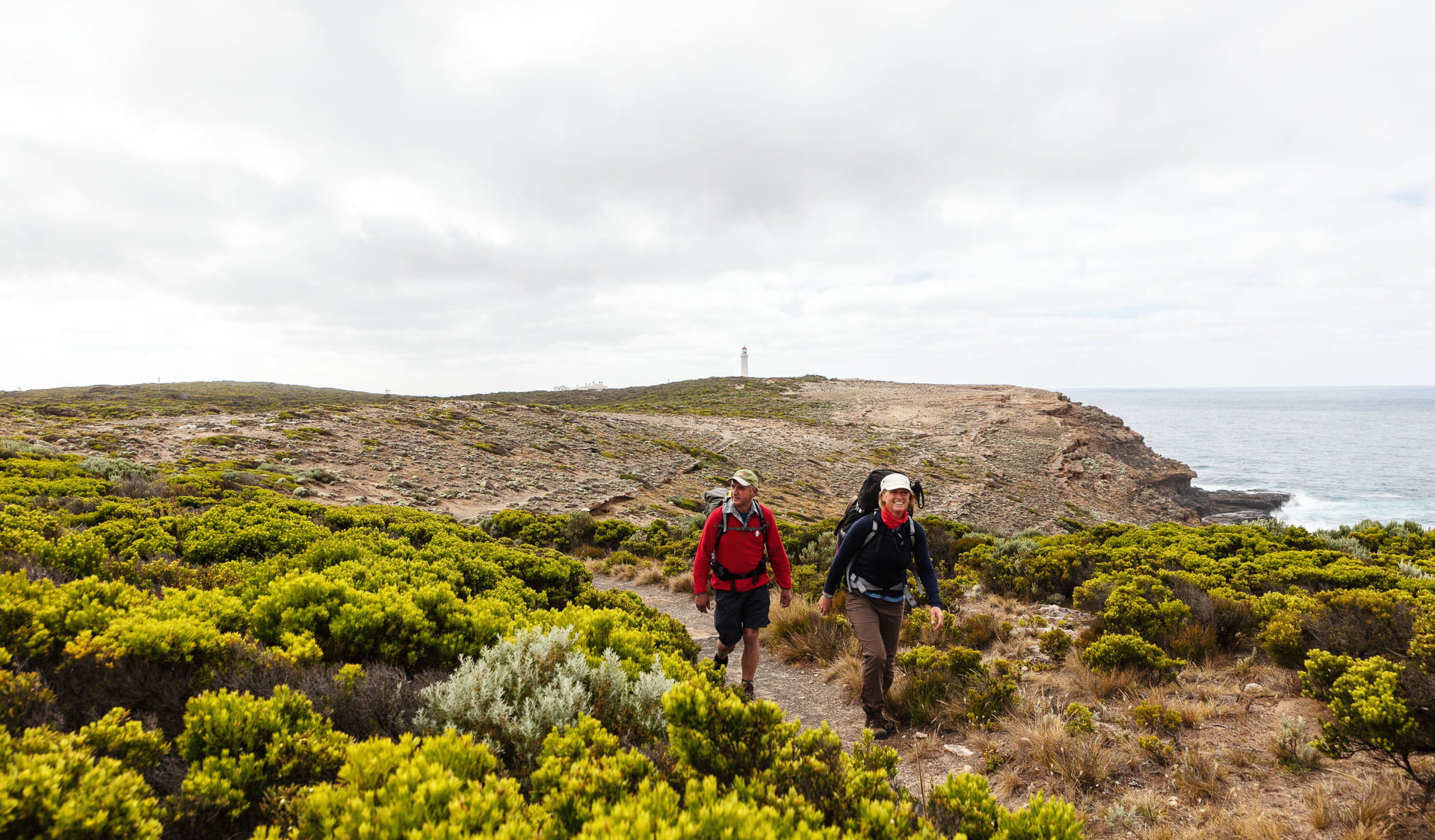 A man and woman hike on a rocky coastline covered with short green shrubbery and blue ocean wave-break in the distance. 