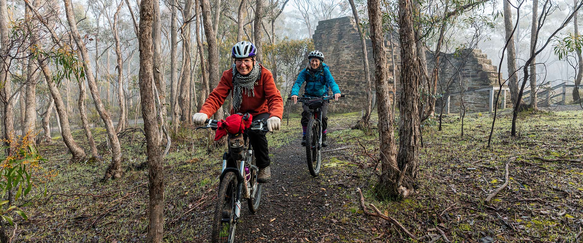 Two females ride mountain bikes past the remnants of a historic structure built from large stones