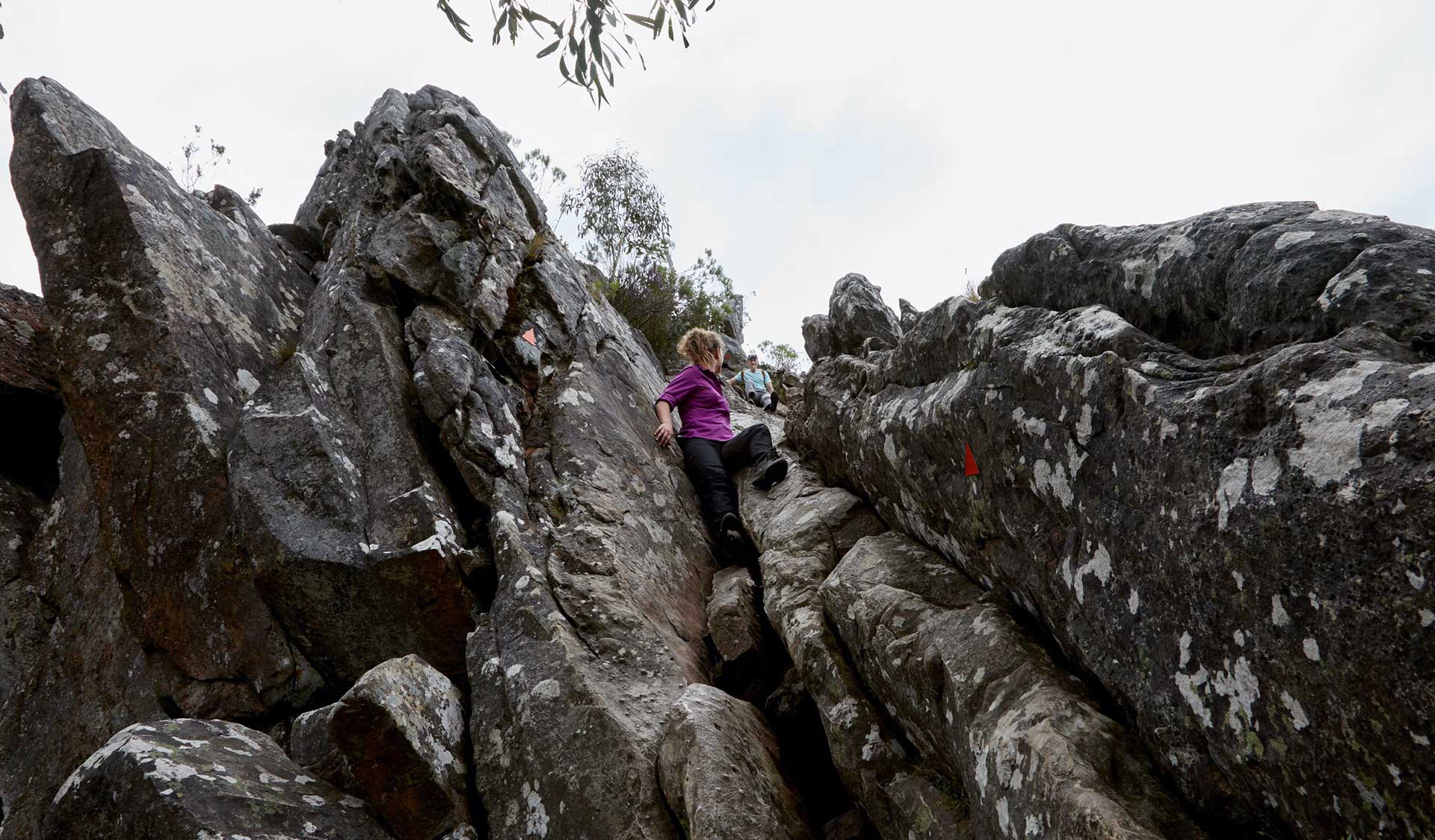 A woman in a purple shirt scrambles up through rocky terrain along the path.