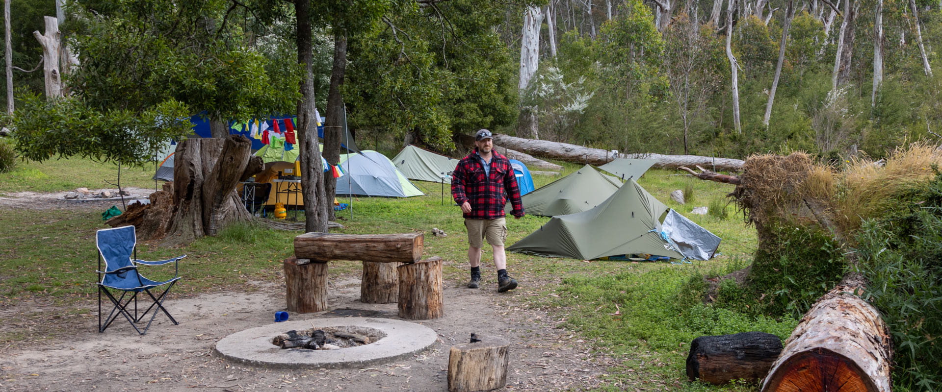 A man walks past a fire pit and rustic wooden log bench, with tents pitched in the campsite behind him bordered by large trees and dense vegetation.