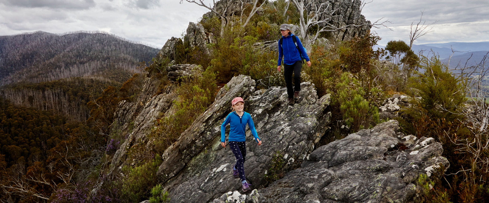 A woman and young girl stand atop a rocky peak to admire the mountain views surrounding them.