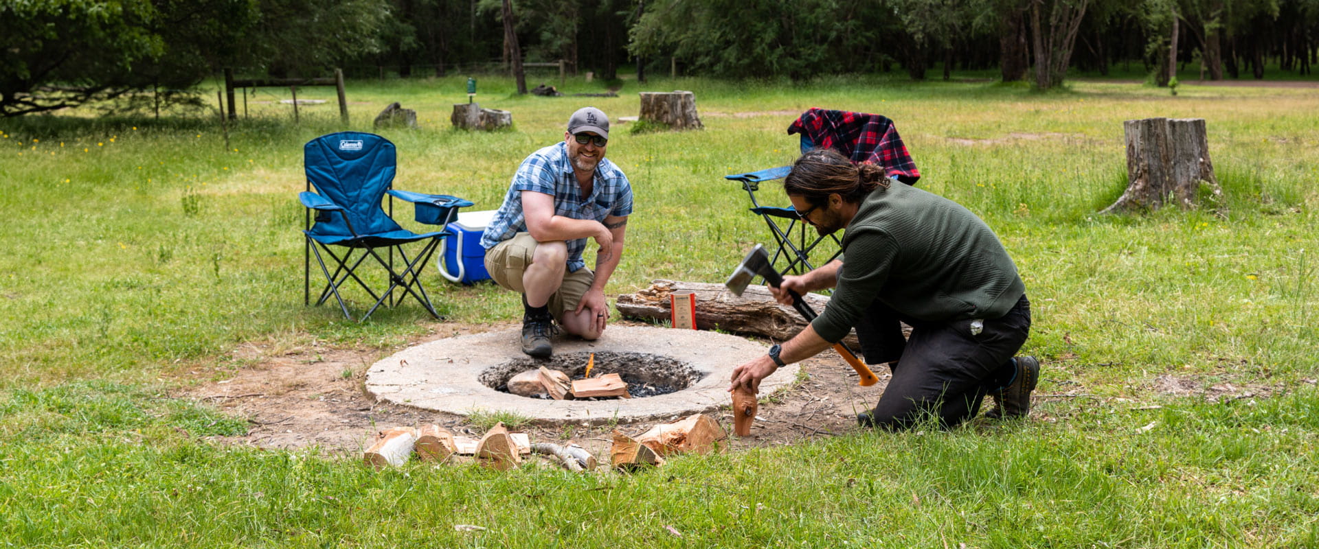 A man kneels to chop firewood while another man helps to tend to the fire pit nestled in front of their two camping chairs and esky.