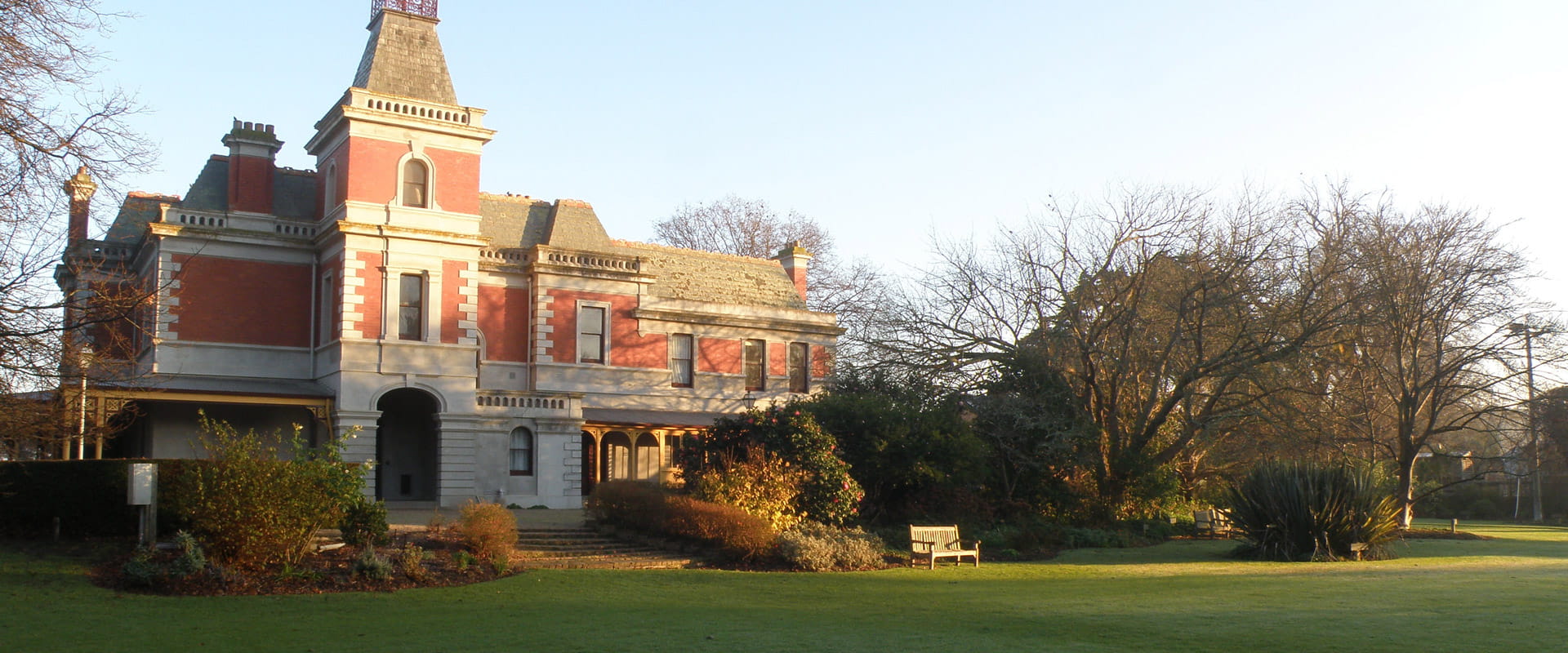A wooden garden bench sits on a manicured grass lawn leading up to the steps of a multi-storey historic heritage building with a red and white facade.