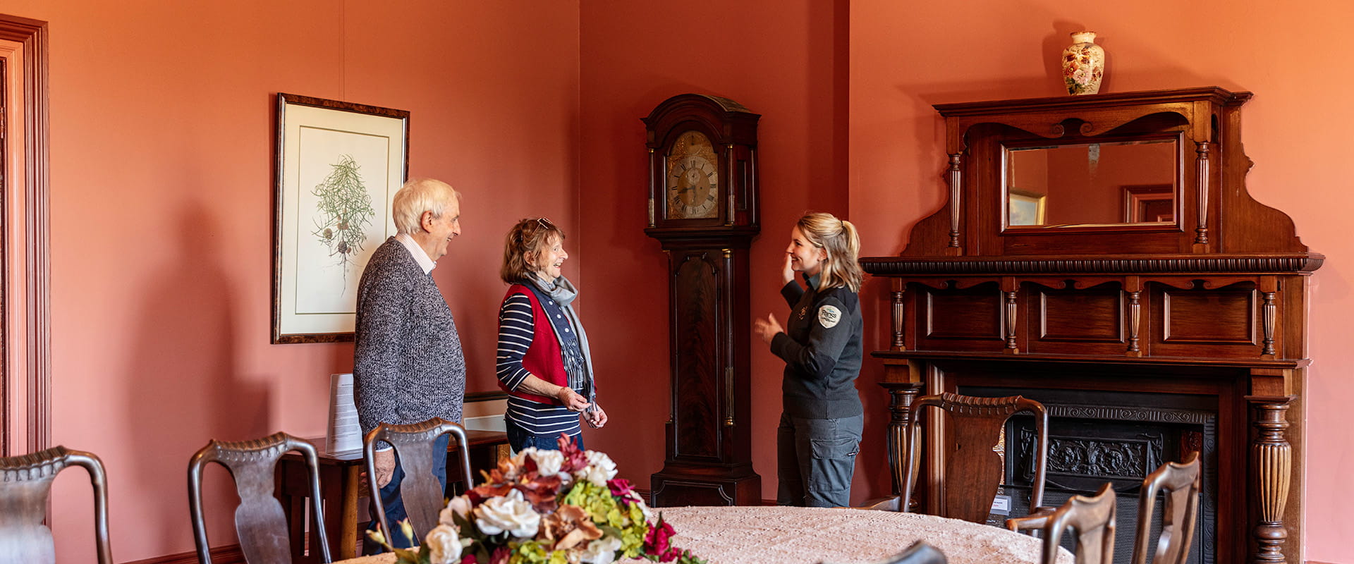 A park ranger points to an old grandfather clock while giving a tour to a man and woman in the dining room of a heritage building with high ceilings and salmon-coloured walls.