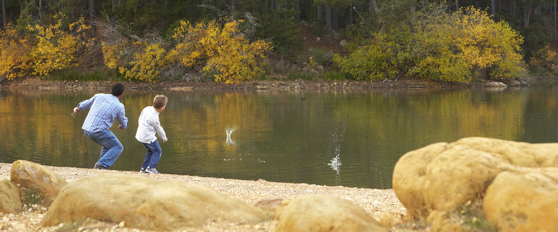 A father and son skip stones across a reservoir from a pebbly beach.