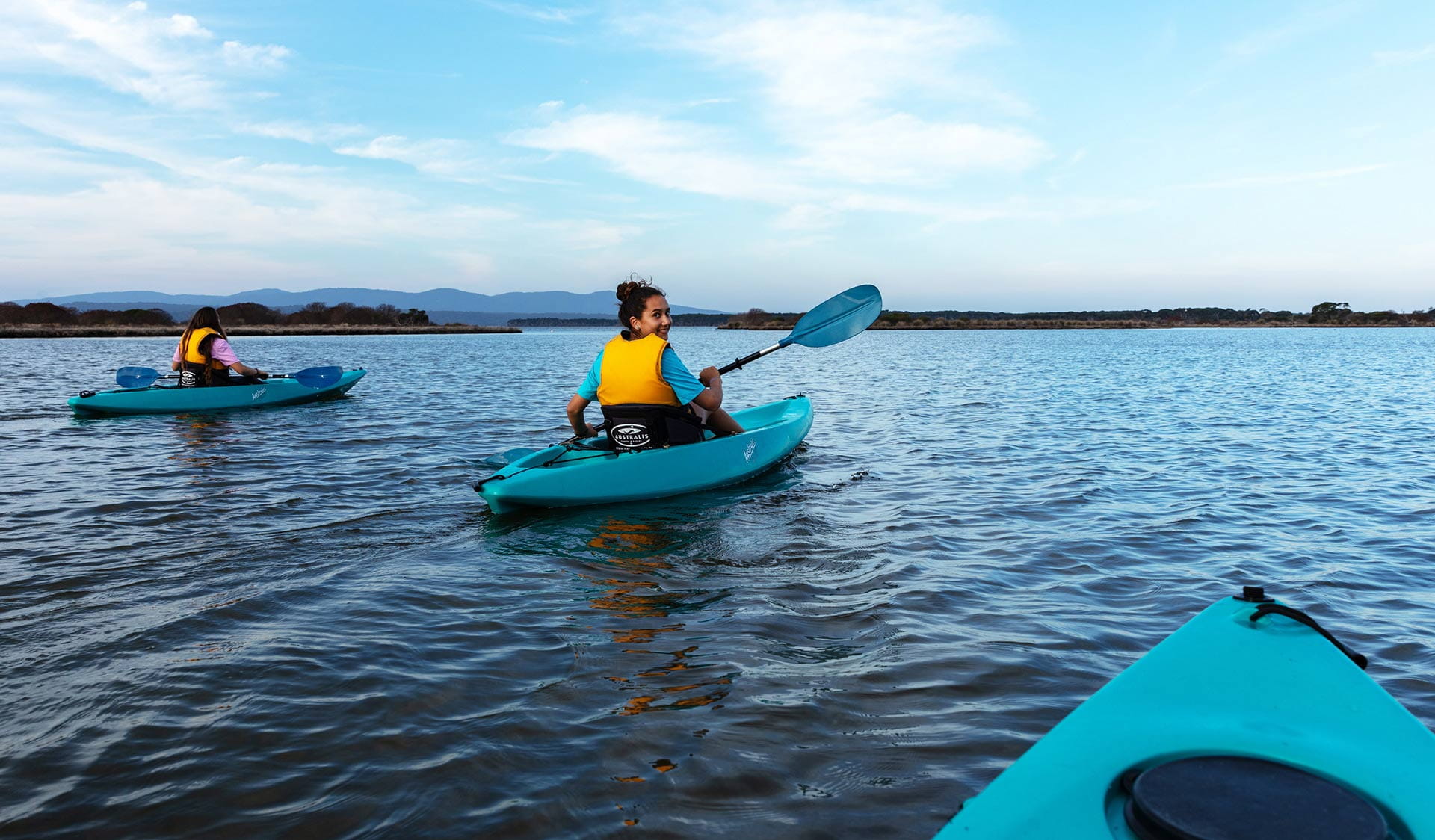Two teenage girls kayaking on Mallacoota Inlet with mountains in the background.