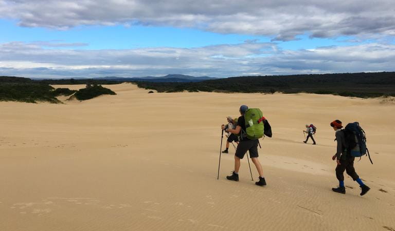 Four friends walk across the sand dunes along the Wildernous Coast Walk close to Mallacoota Inlet.