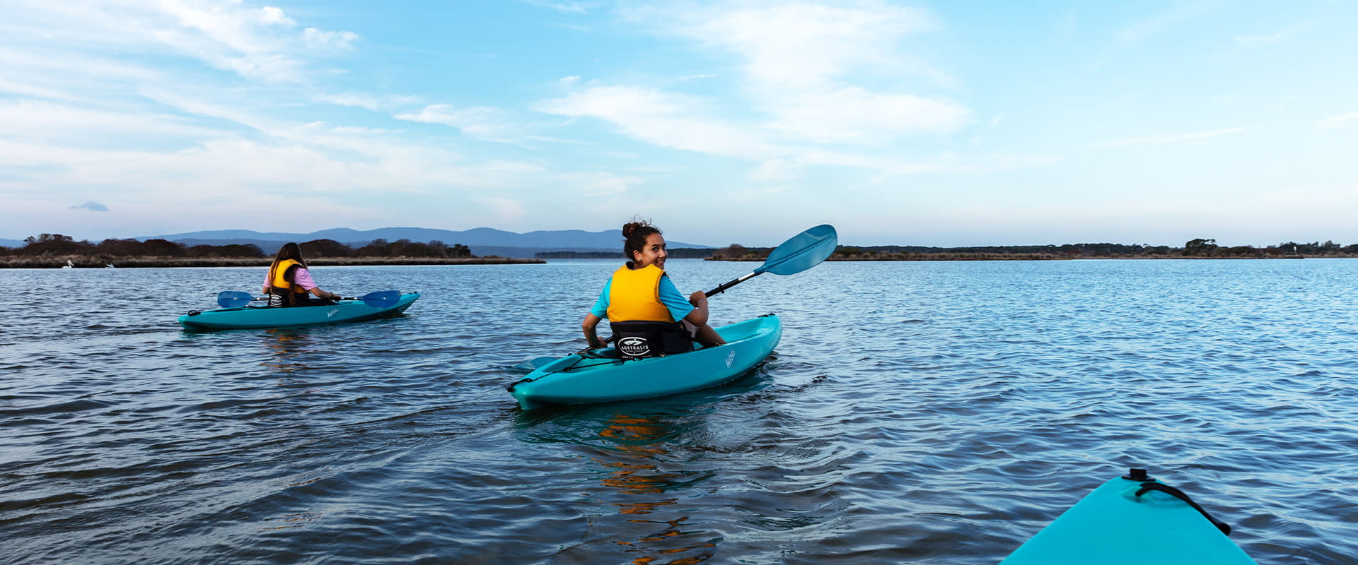 Two young women paddle blue kayaks on a large body of water with mountains in the background