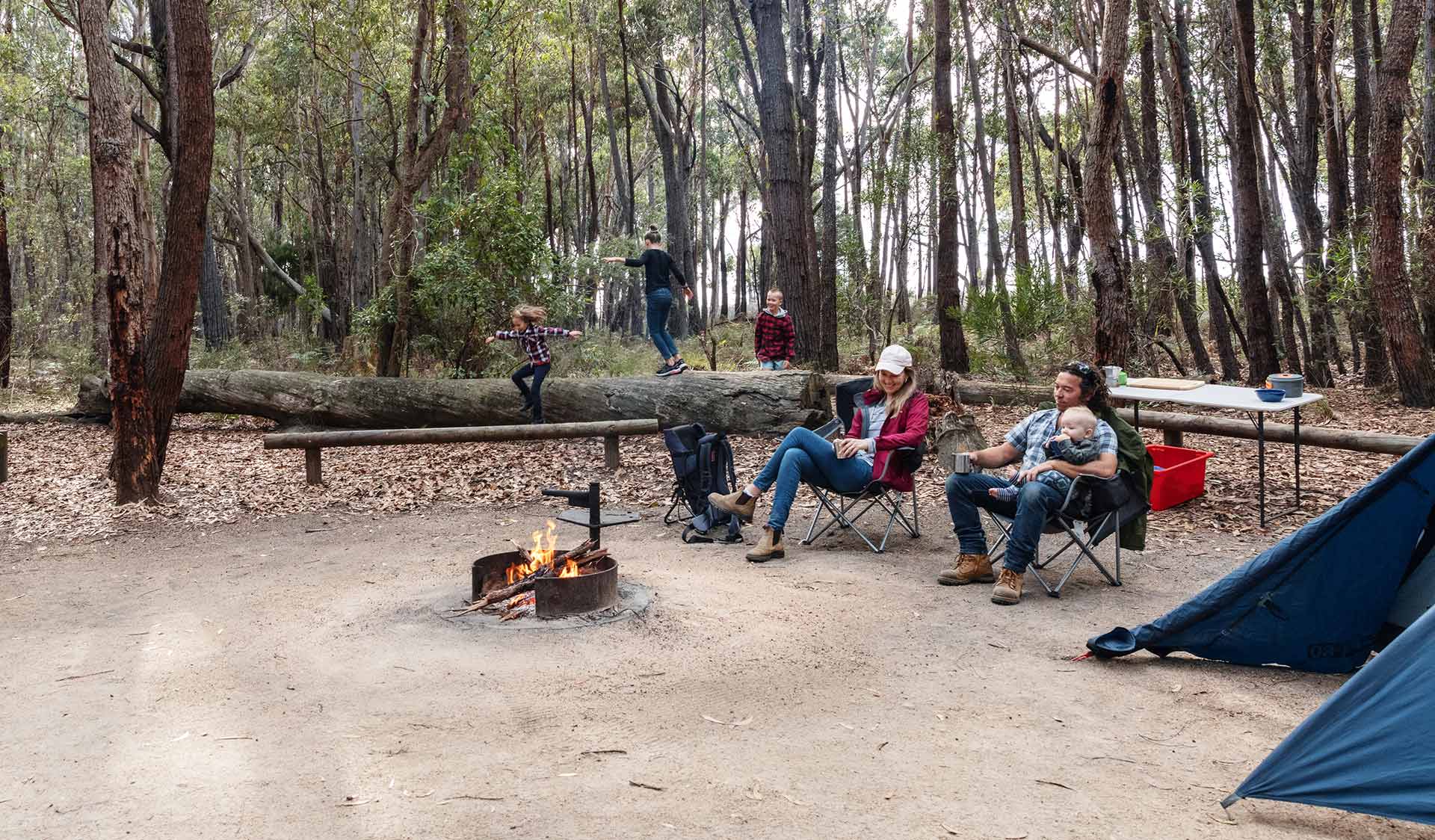 Mum and Dad sit around the campfire with their infant son while their three other children play in the background.