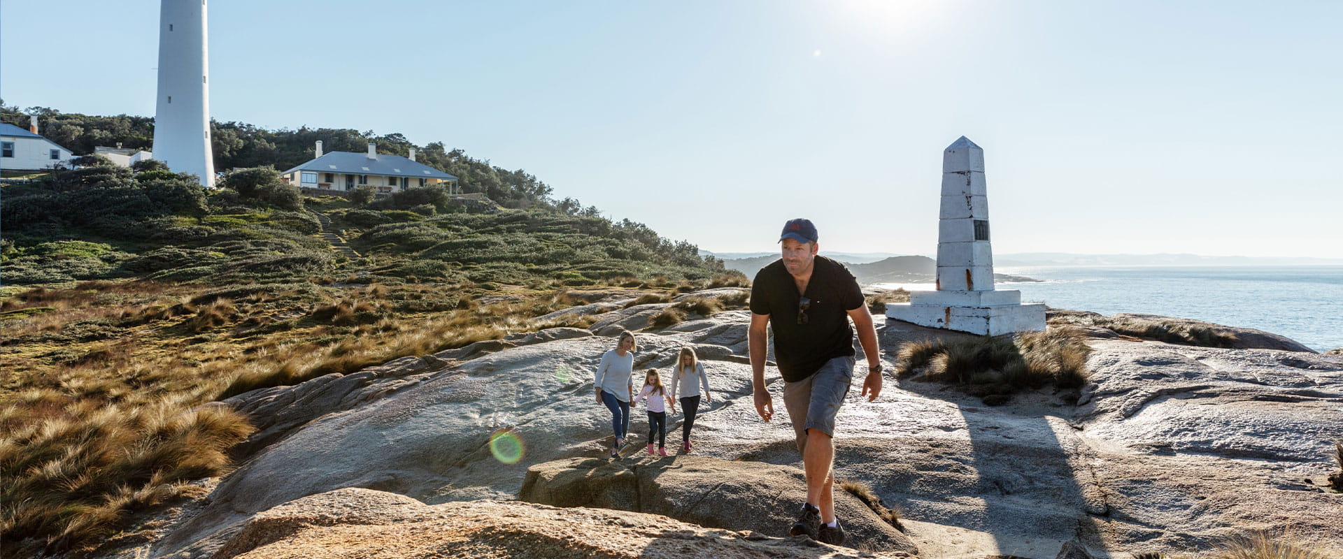A father walks over rock slabs with his young family in the background in front of a memorial statue and lightstation in the background