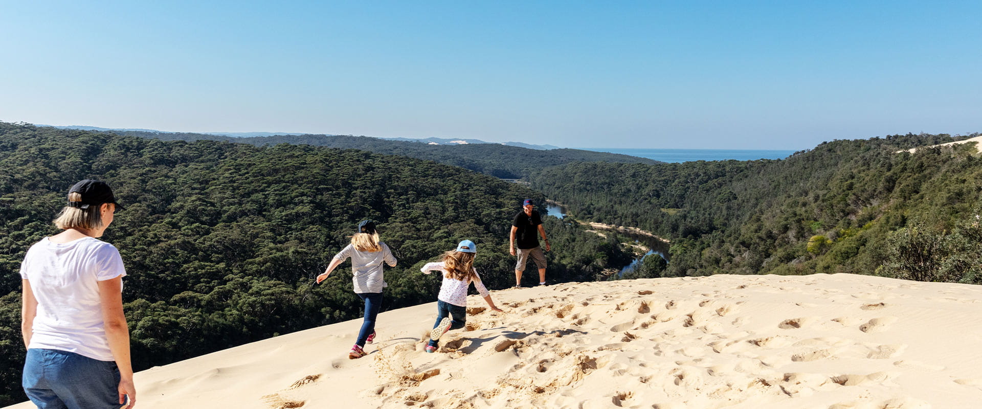 Two young girls run towards their dad on a sand dune above a river and the ocean far in the background