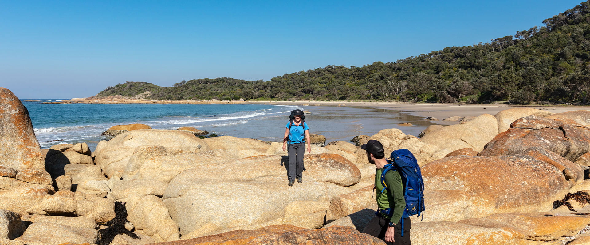 A man waits for his female hiking partner as they walk through a pile of rocks at one end of a sandy bay with the water in the background