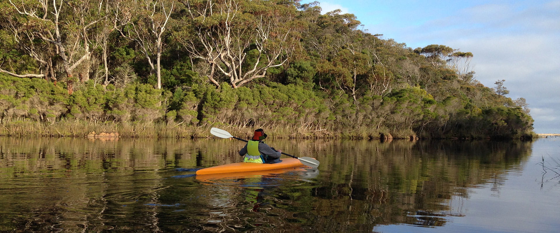 A woman wearing a red bucket hat paddles an orange kayak on still water