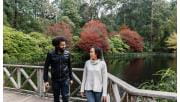 A man with an afro wearing a leather jacket and woman wearing a cream knitted jumper turn and walk away from a lake in the Dandenong Ranges Botanical Gardens.