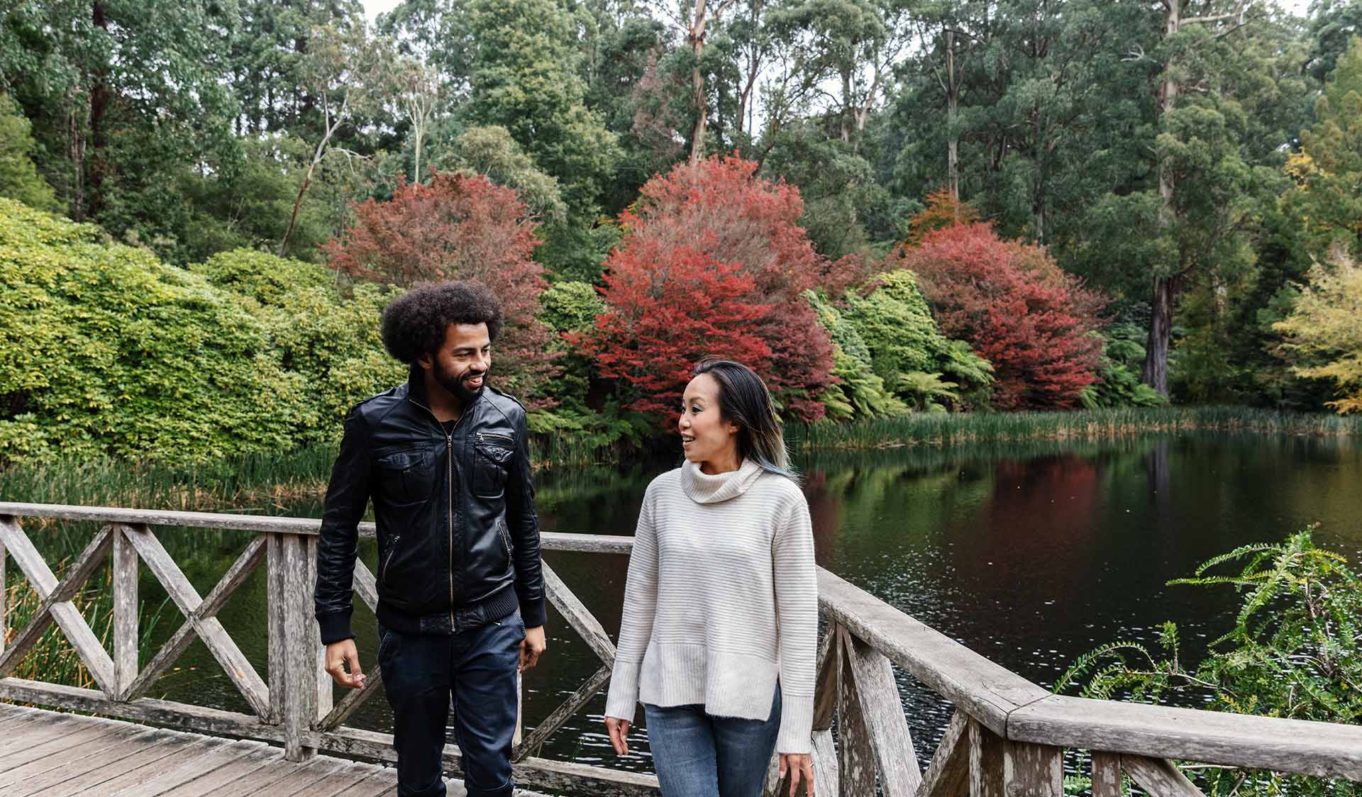 A man with an afro wearing a leather jacket and woman wearing a cream knitted jumper turn and walk away from a lake in the Dandenong Ranges Botanical Gardens.