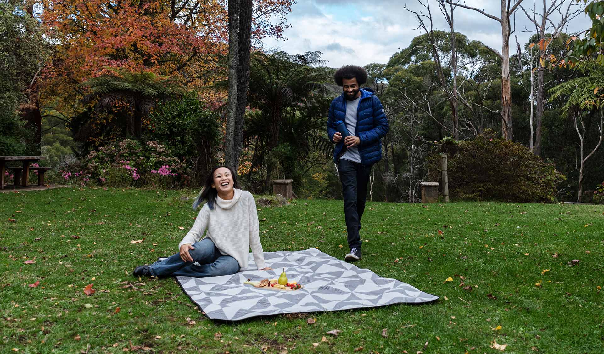 A man in a blue puffer jack walks towards a women with black hair wearing a cream jumper to join a picnic.