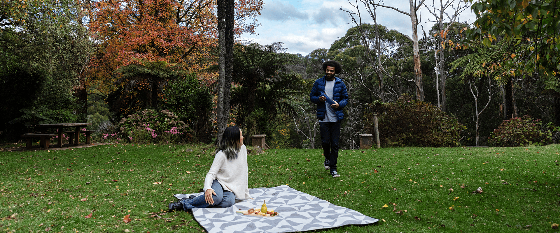 A couple enjoying a picnic of cheese and fruit on a rug on grass, a tall tree fern sits behind them and deciduous trees in autumn colour.