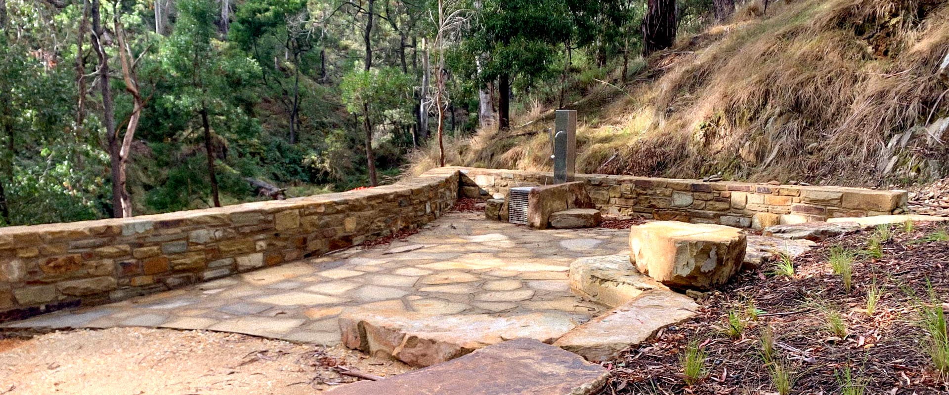 A mineral spring fountain surrounded by brickwork sits amongst a hilly, rugged bushland.