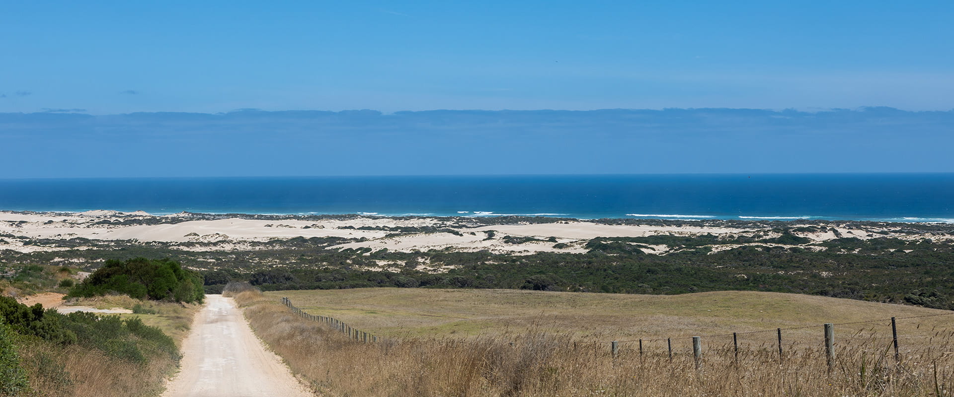 A straight dirt road leads down into sand dunes beyond which lies the ocean