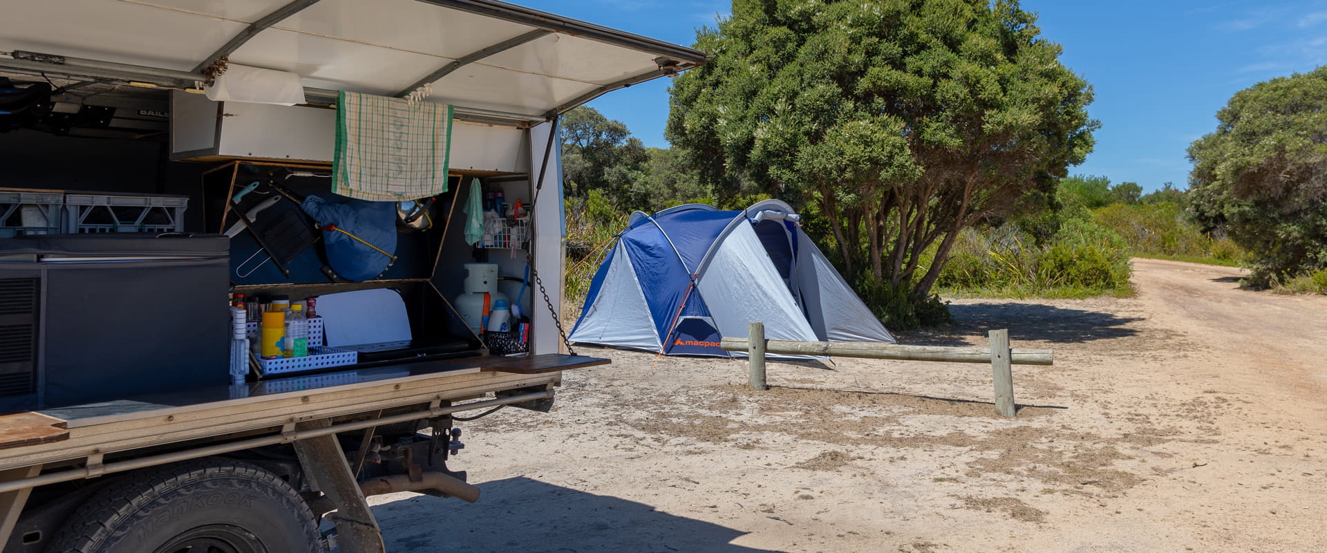 A camping trailer sits open in front of a tent pitched under a tree in a beachside campground.