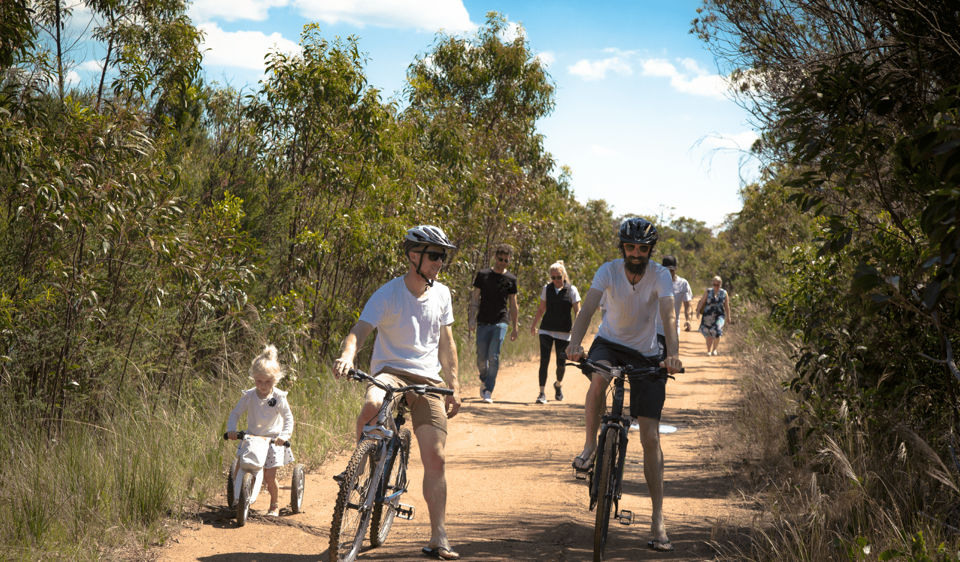 Two men and a female toddler ride bikes in front of people walking at Frankston Nature Conservation Reserve