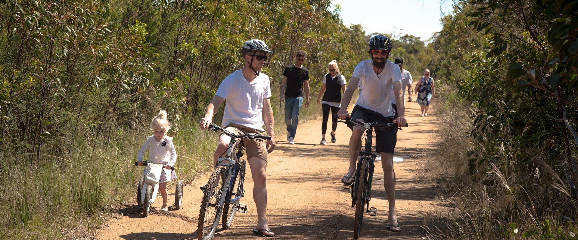 A group a people ride bikes and walk along a trail surrounded by native bushlands