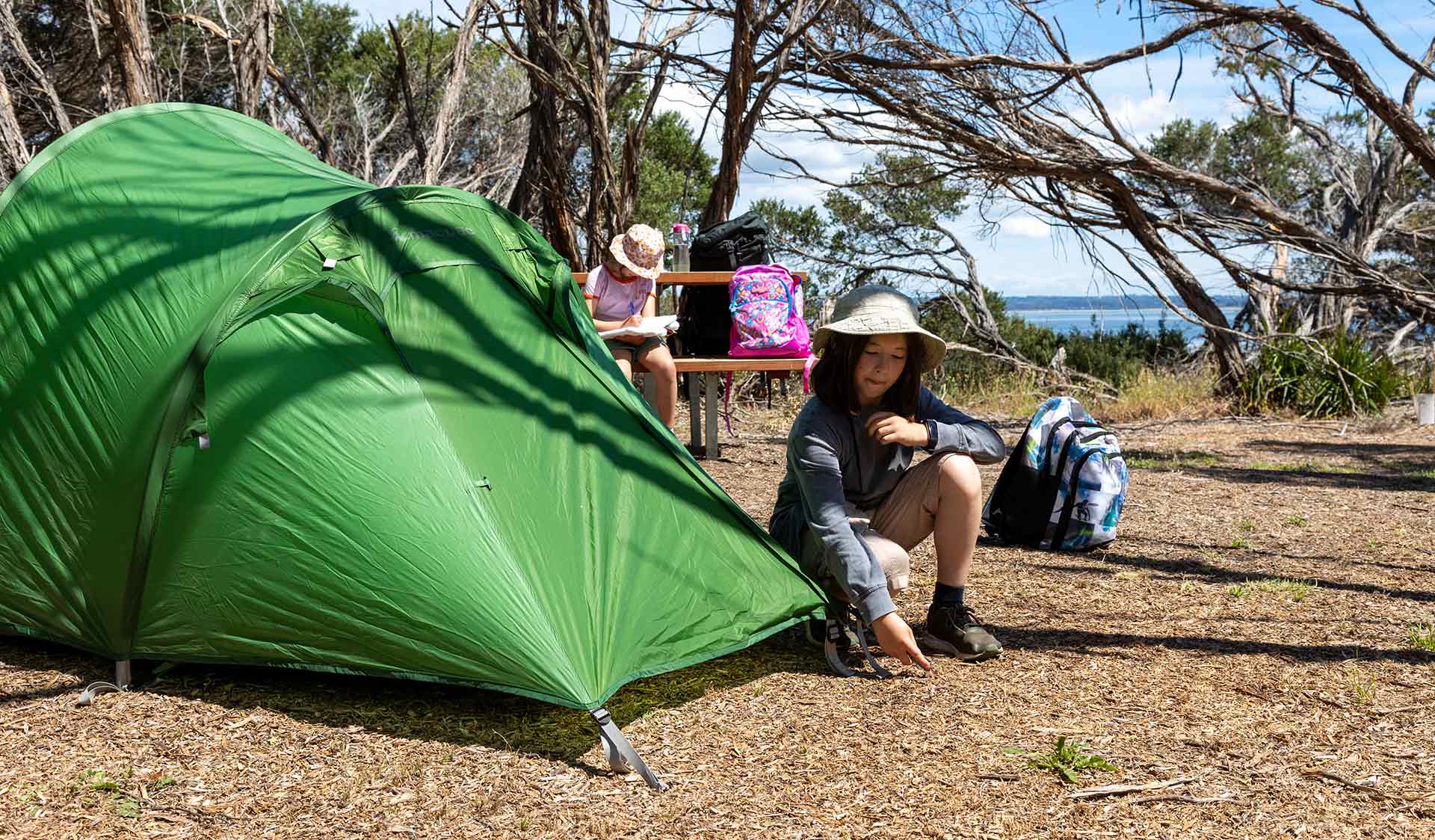 A young boy finishes setting up his tent while his sister sits at a picnic table with views of Western Port in the background at Fairhaven Campground at French Island National Park 
