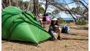A young boy finishes setting up his tent while his sister sits at a picnic table with views of Western Port in the background at Fairhaven Campground at French Island National Park 