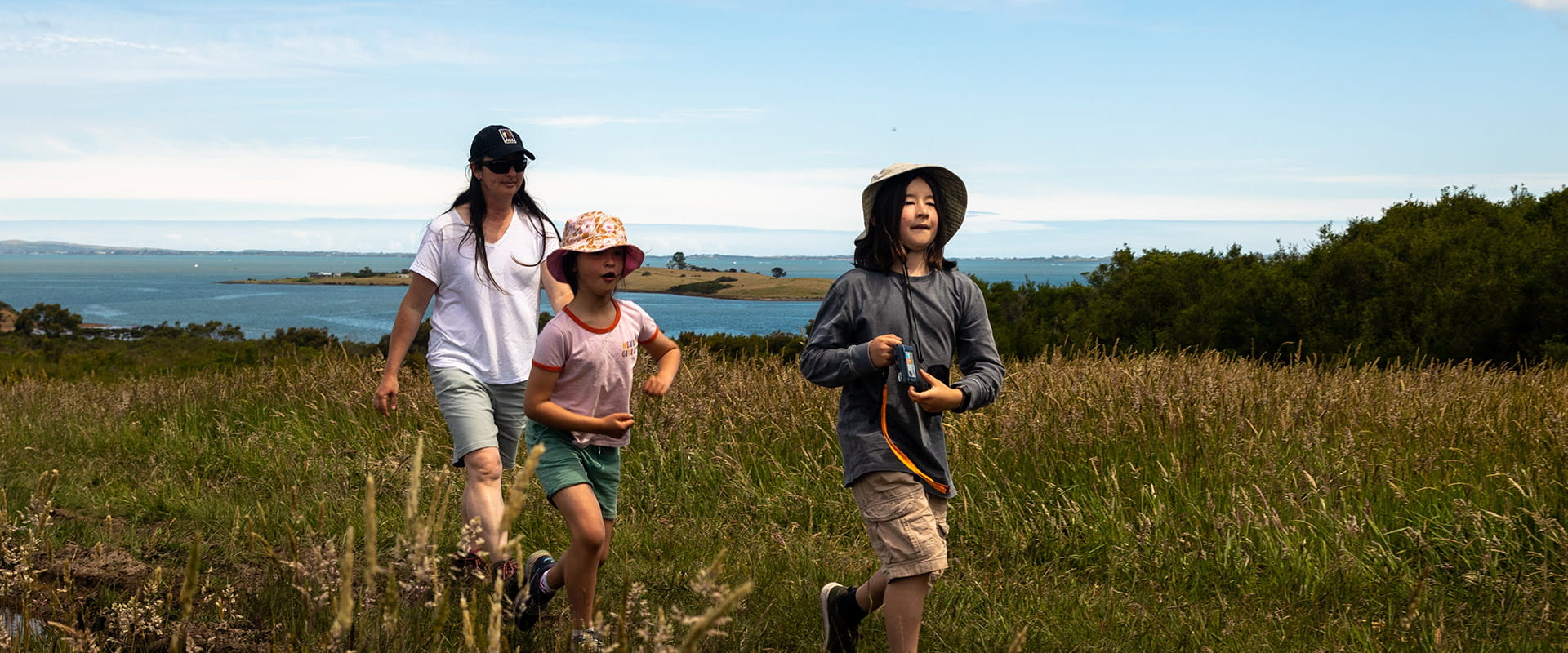 A mother and two children walk along a grassy area on a hill that overlooks the bay