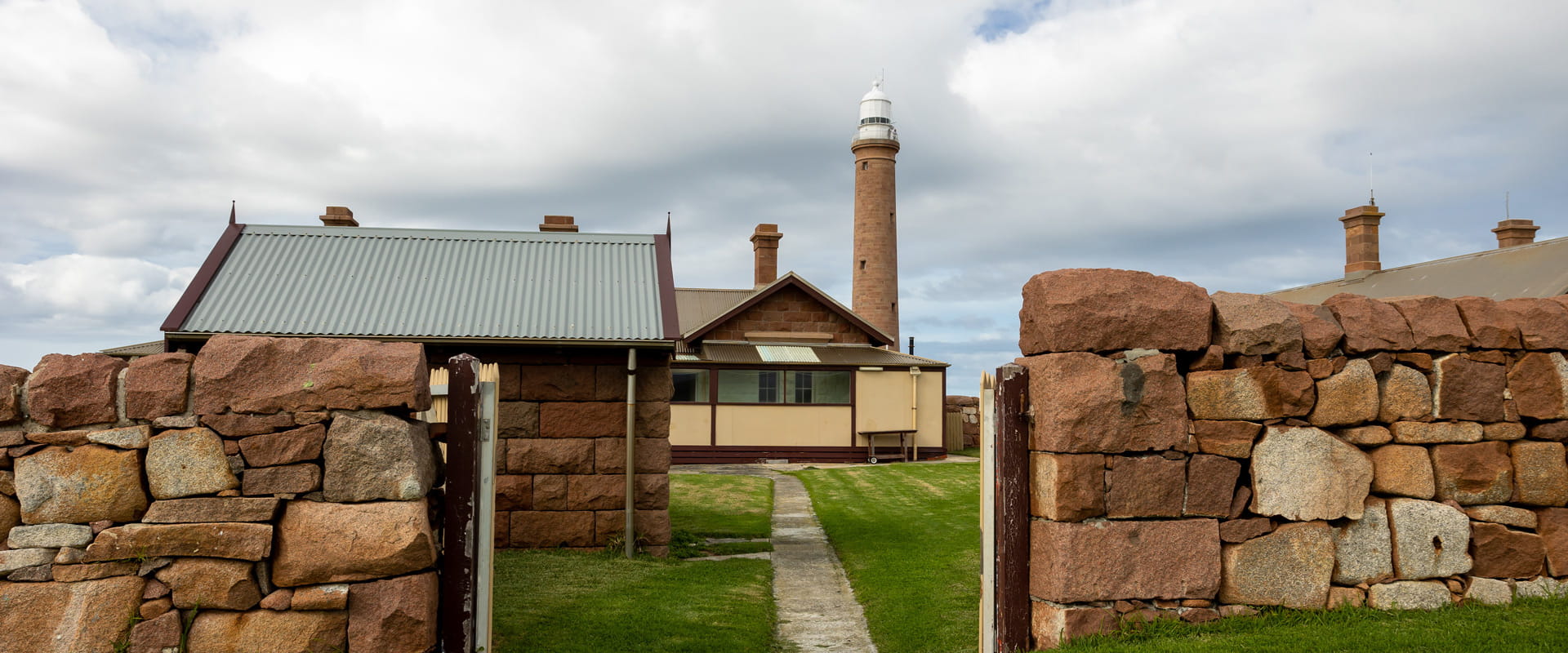 A small path leads up to an old cottage seated beneath a lighthouse, visible through an open wooden gate supported by a rustic rock fence.