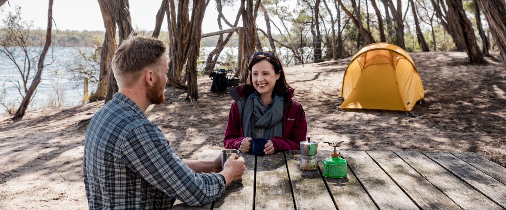 Two people sit at a picnic table talking, a tent behind them in a peaceful forested area. 