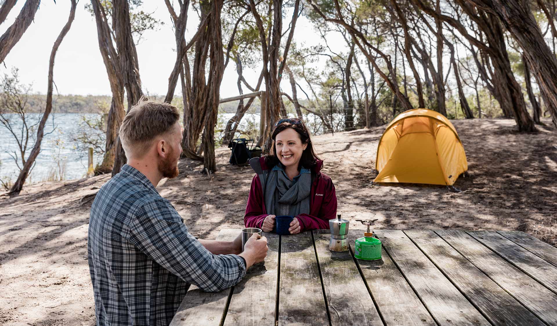 A woman enjoys a cup of tea while sat at a picnic table infront of her tent at Bunga Arm Campsite in the Gippsland Lakes.
