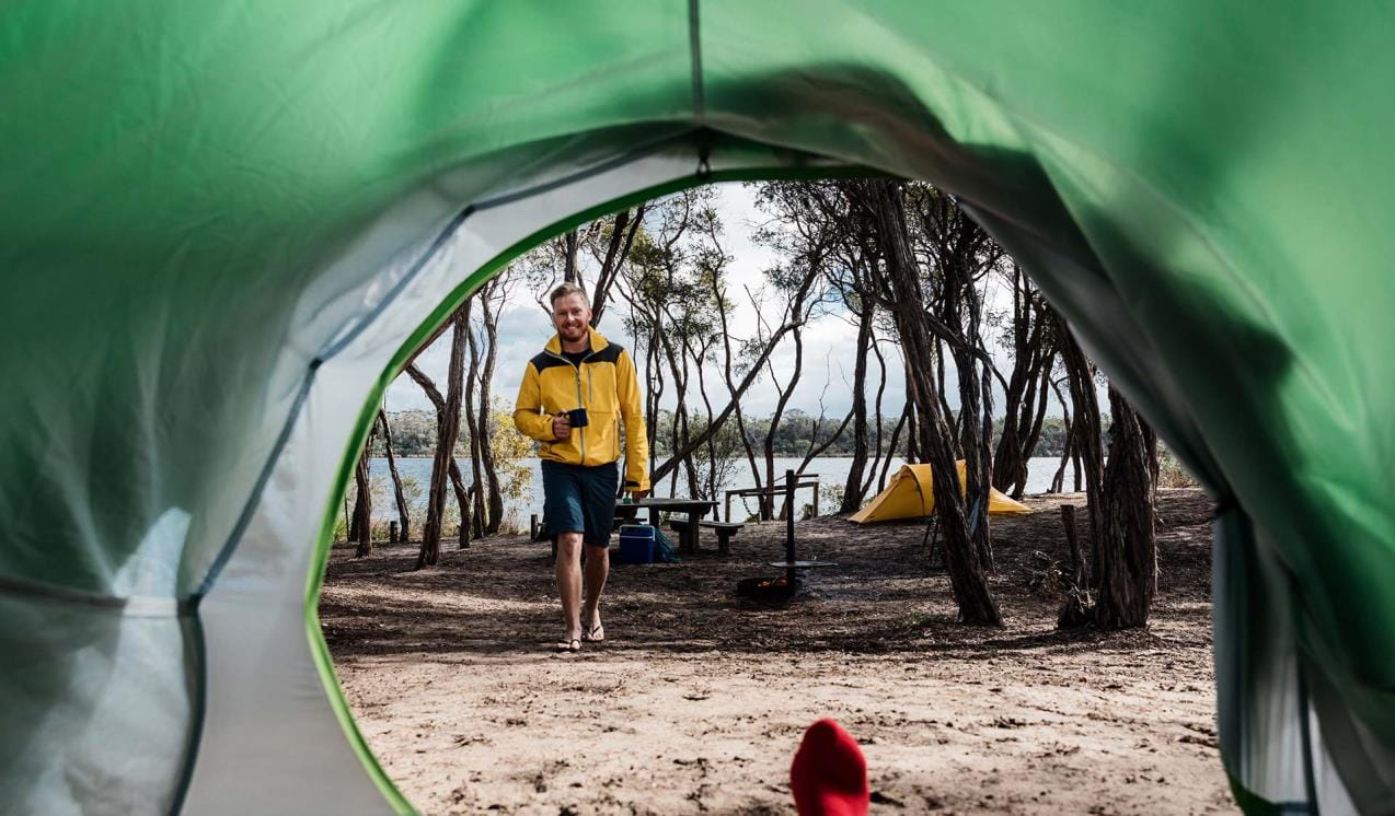 A man in a yellow jacket holding a cup of tea walks towards his partner in a tent 