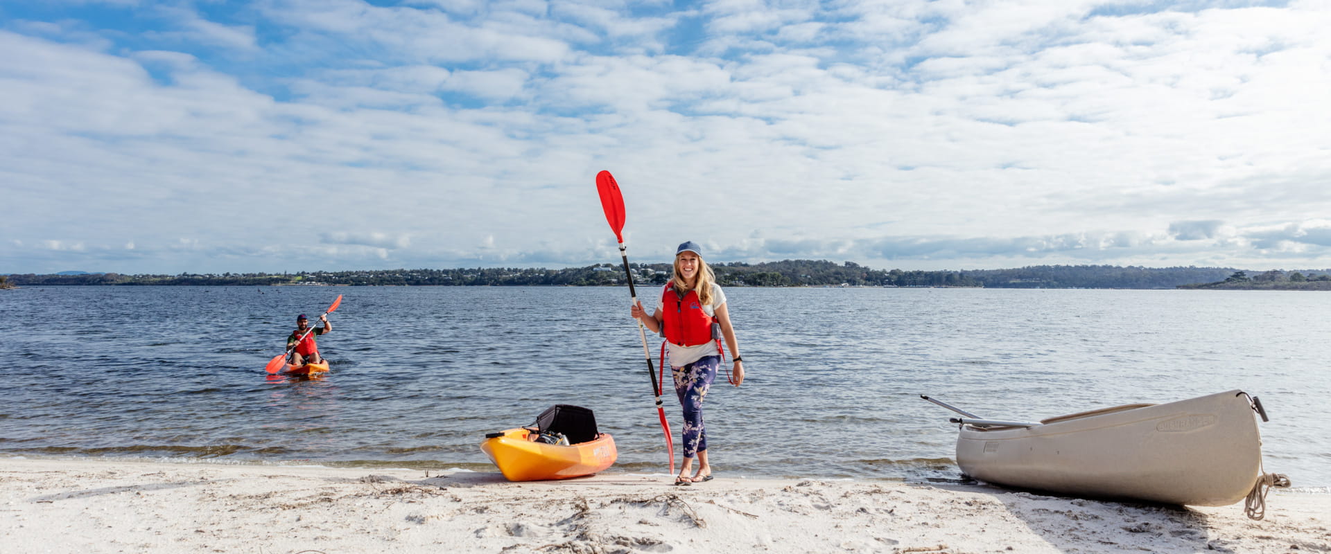A person pulls their kayak out of the water onto an ocean beach. It's a clear sunny day. Someone kayaks in the background. 