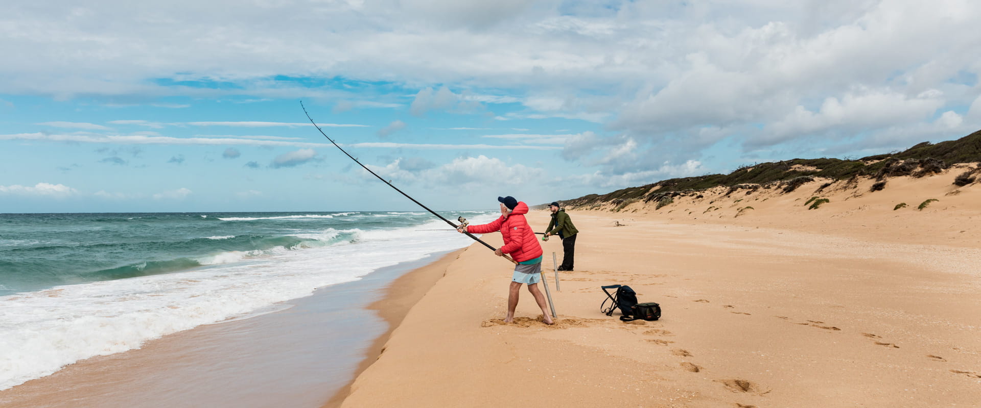 Two people cast fishing lines into the ocean from the edge of the beach. 