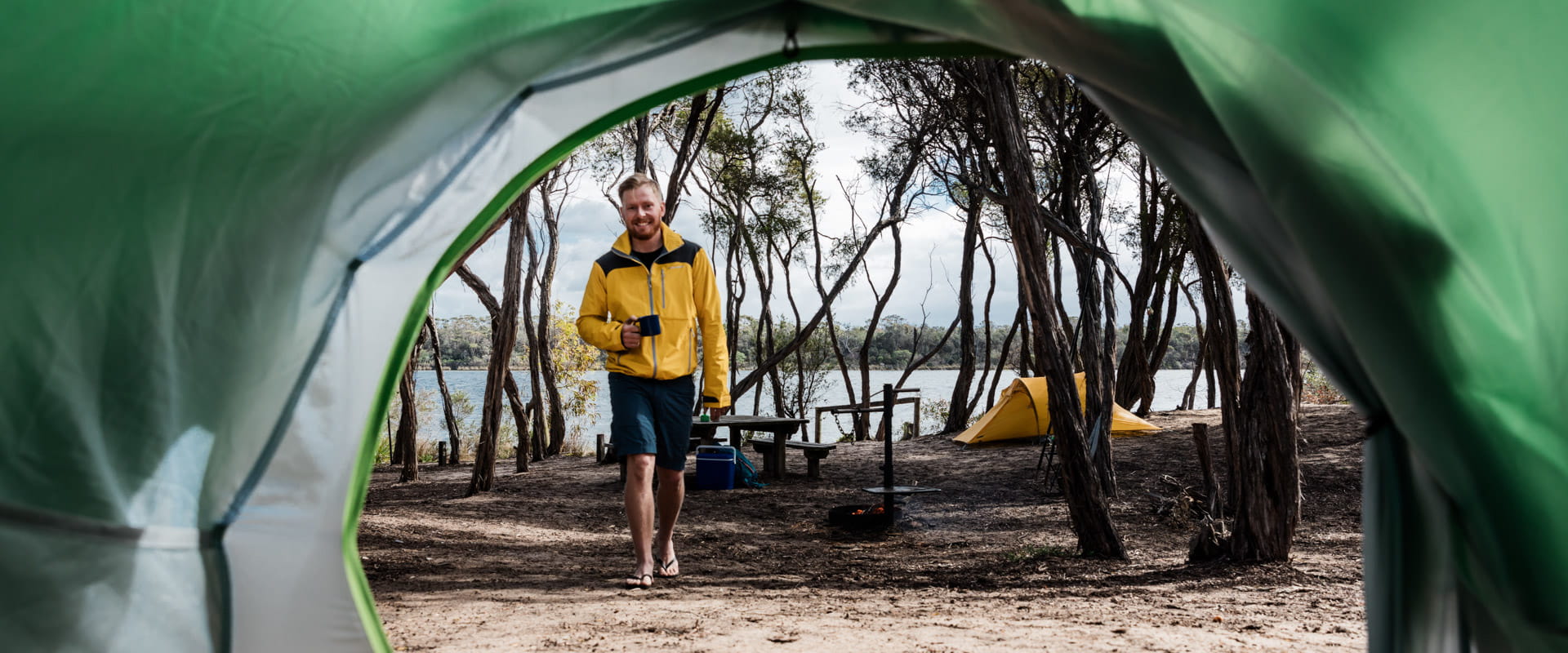 The point of view from inside a green tent looking out to a lake. A man approaches the tent with a coffee. 