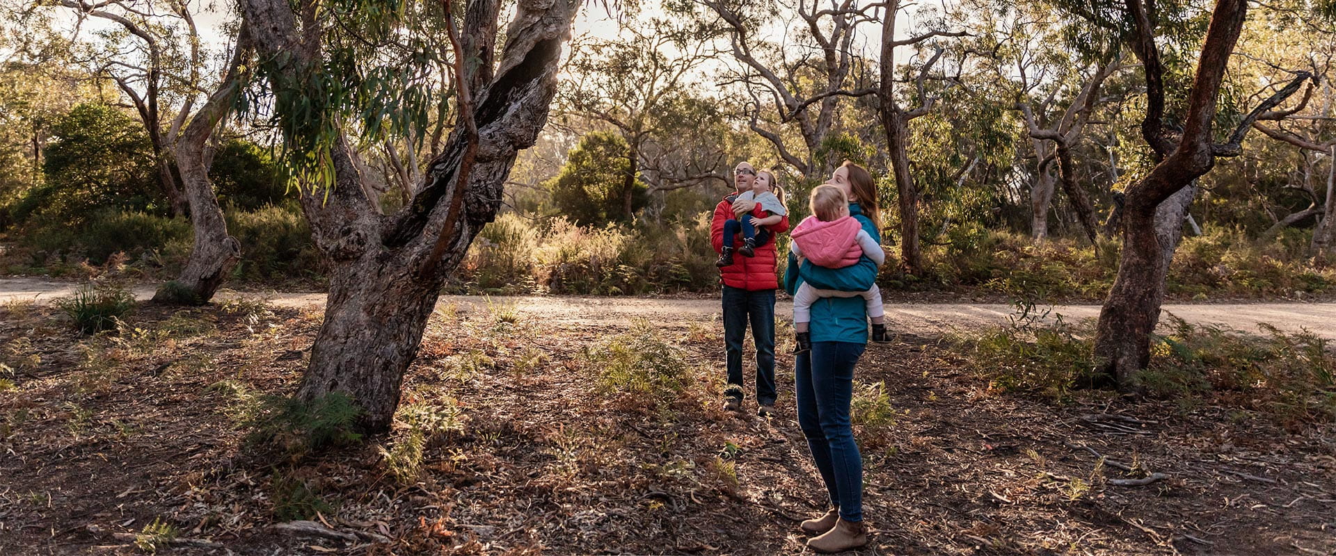 Two adults in warm outdoor clothing standing underneath trees and looking up while holding young children.