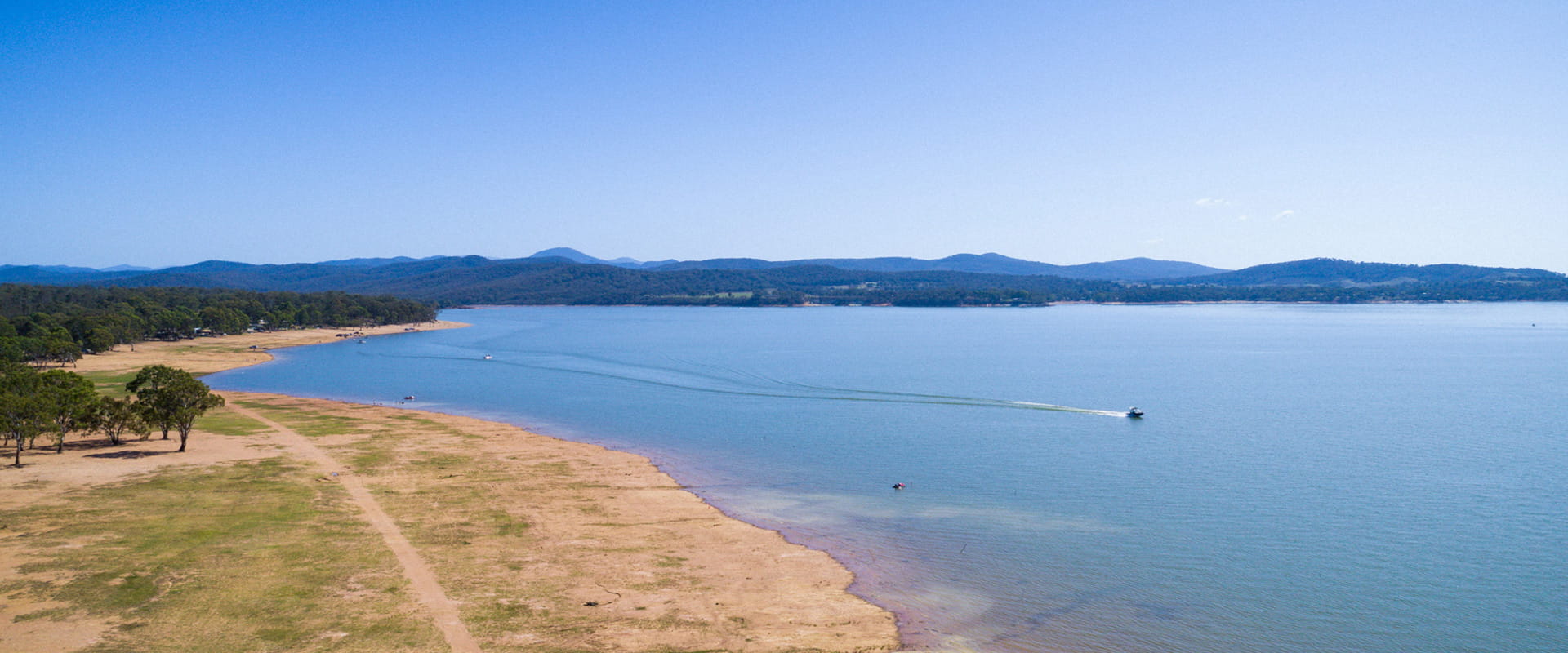 An expansive view of a lake surrounded by green forested hills, a small boat travels along the lake, leaving a white foamy wake behind it.