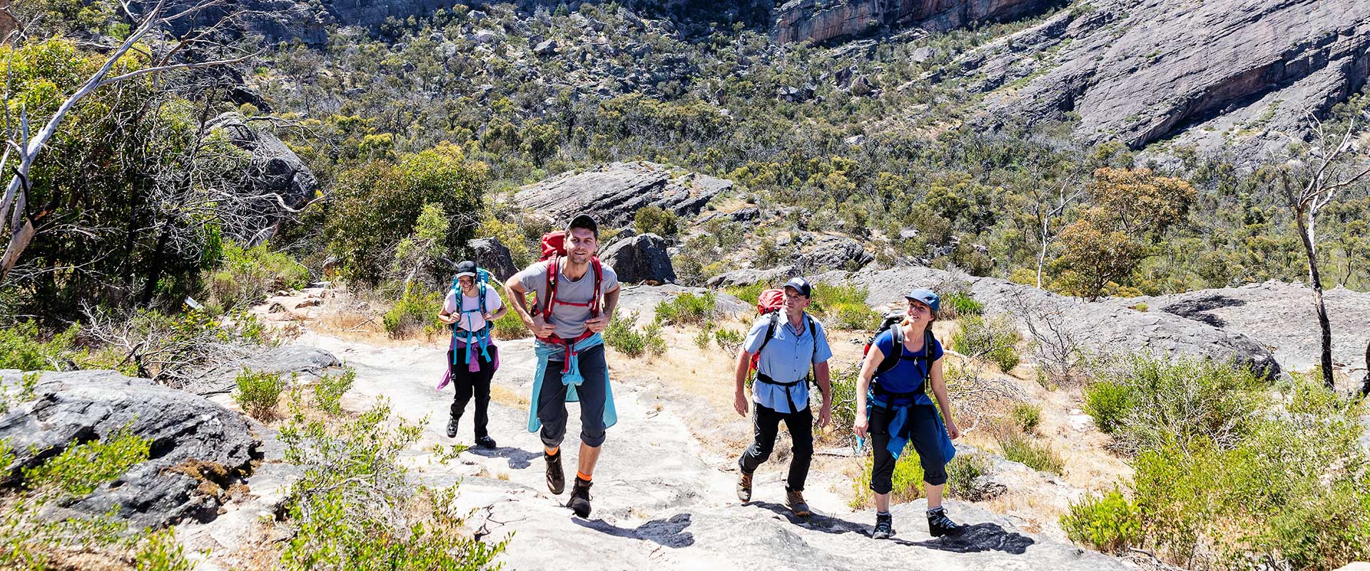 A group of walkers walk along a rocky trail with large cliffs in the background