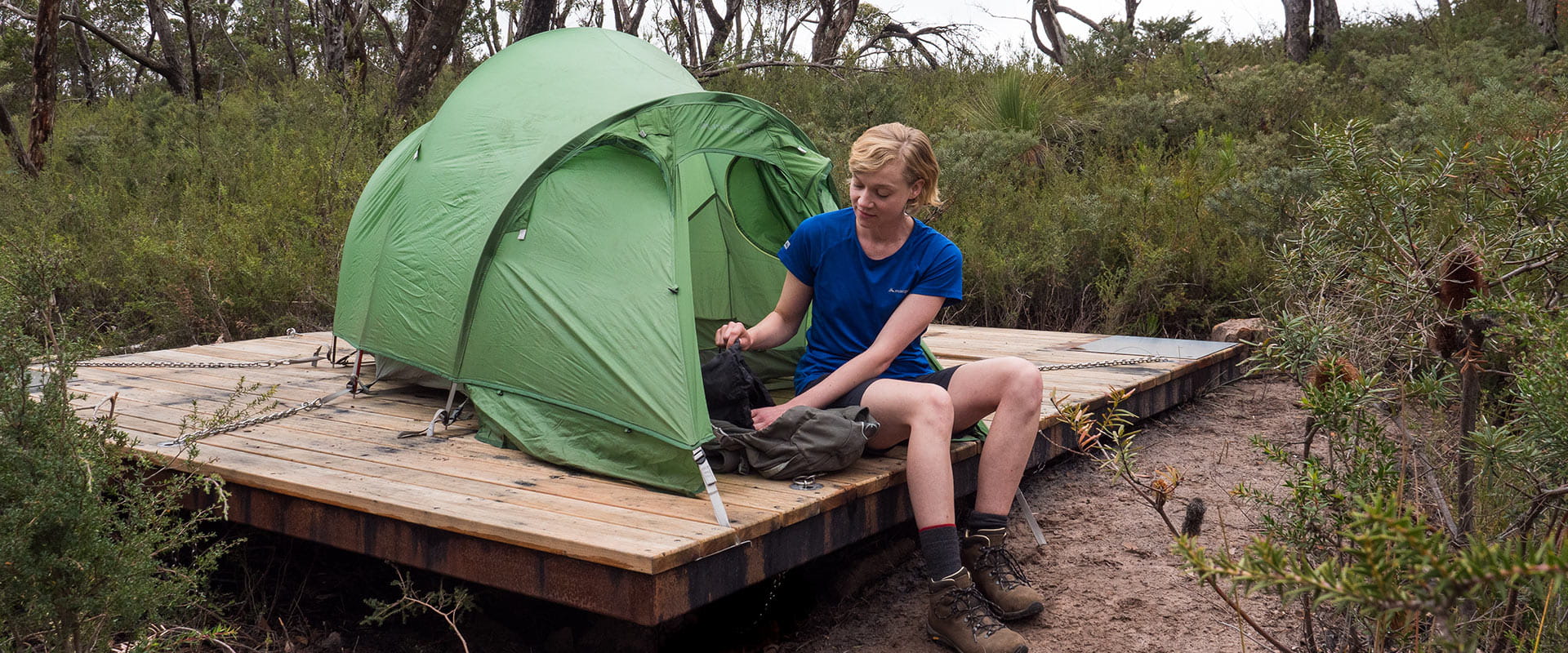 A hiker sits on a tent pad setting up the tent and surrounded by short native foliage