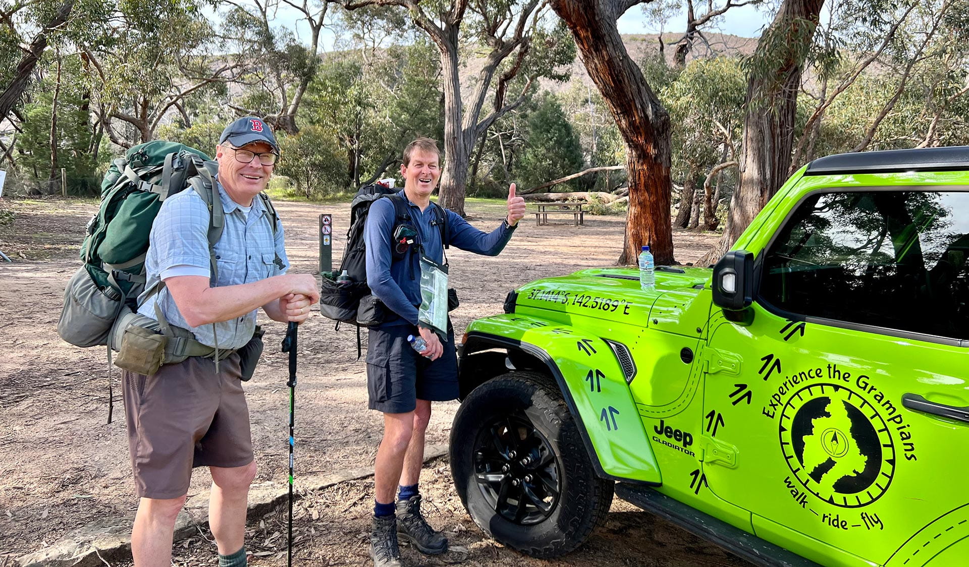 Two hikers with their gear standing and smiling next to a Experience the Grampians branded vehicle.