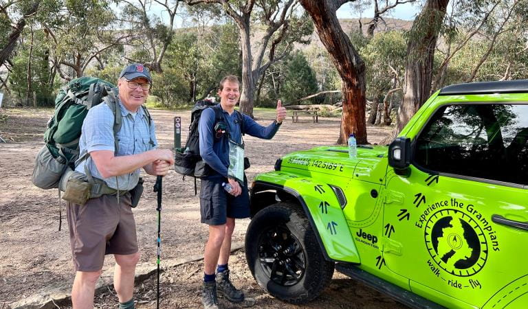Two hikers with their gear standing and smiling next to a Experience the Grampians branded vehicle.