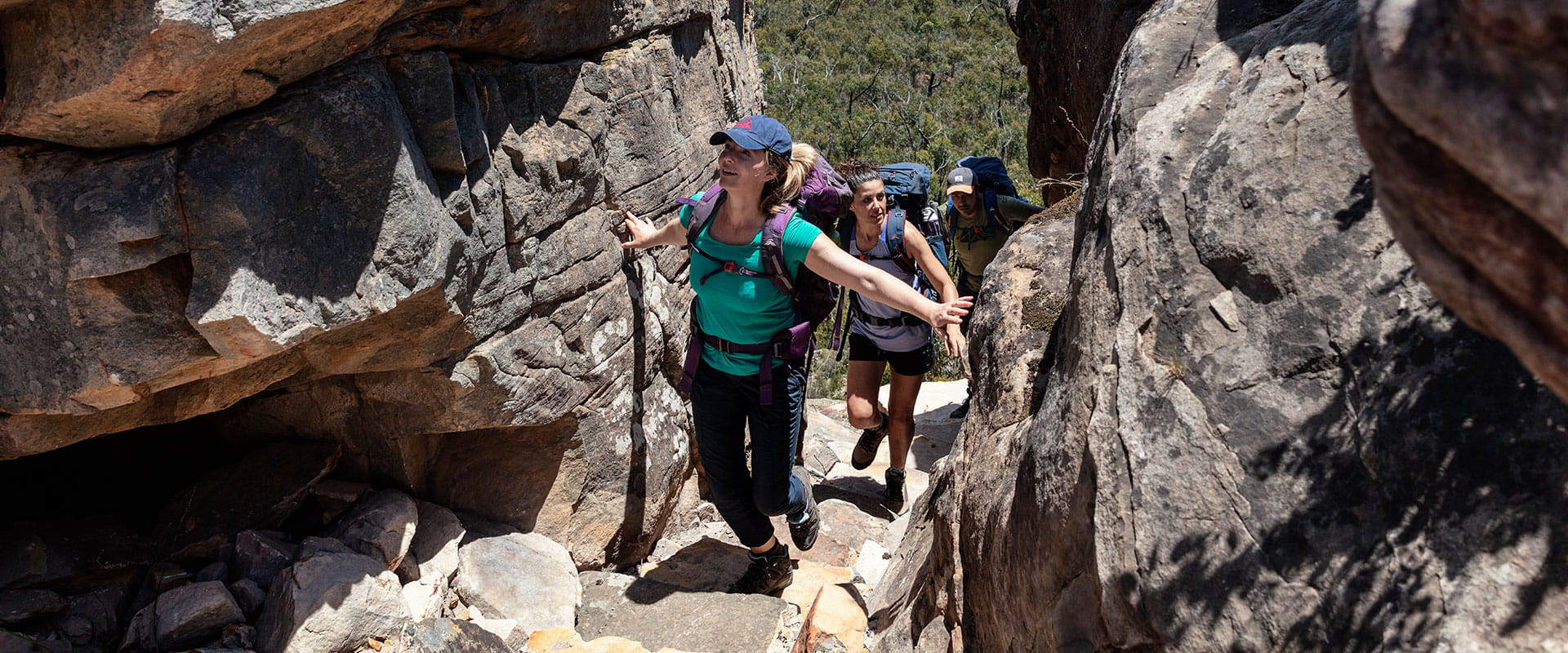 A group of hikers walk through a narrow trail with large stone on either side and a mountainous view in the background 