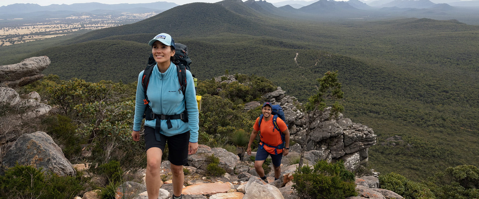 Two hikers ascend a steep trail with mountainous views in the background