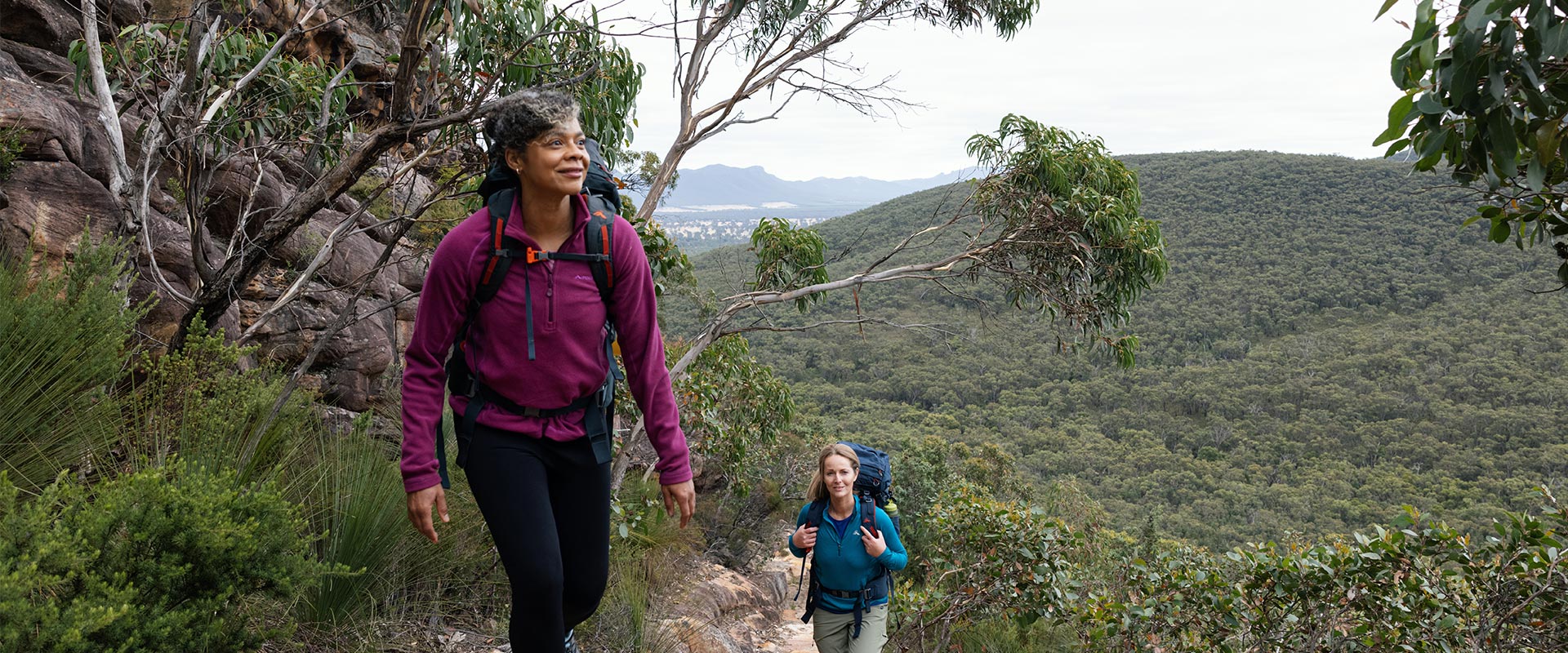 Two hikers ascend the lower slopes along a rocky trail surrounded by nearby hills and dense trees
