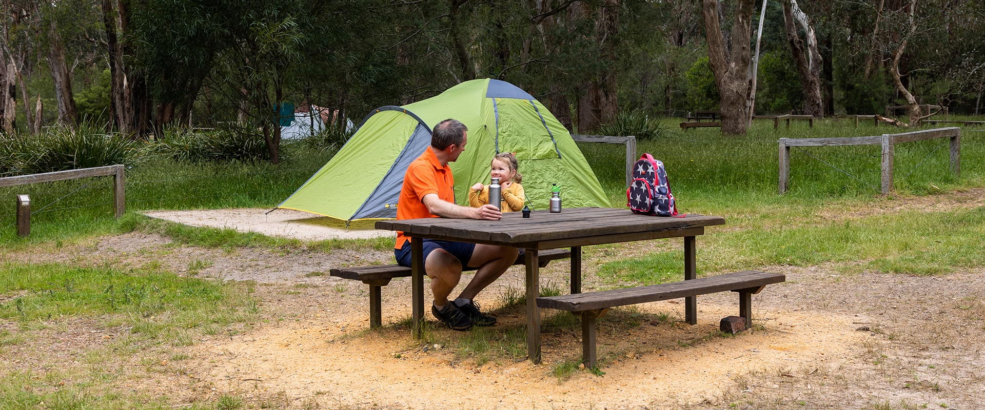 A father and daughter enjoying a drink at a picnic table next to their tent in a grassy campground