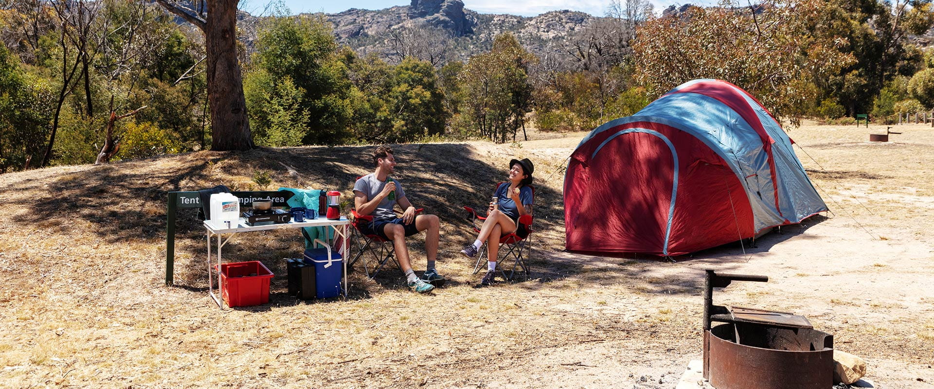 A couple sit in the shade next to their tent with mountains in the background