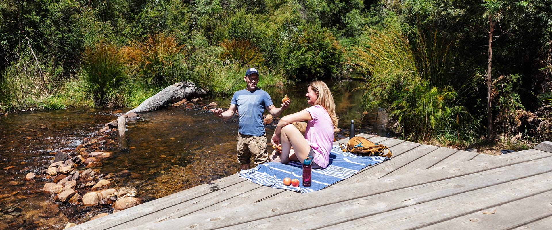 A couple cool themselves by a river and enjoy some refreshments