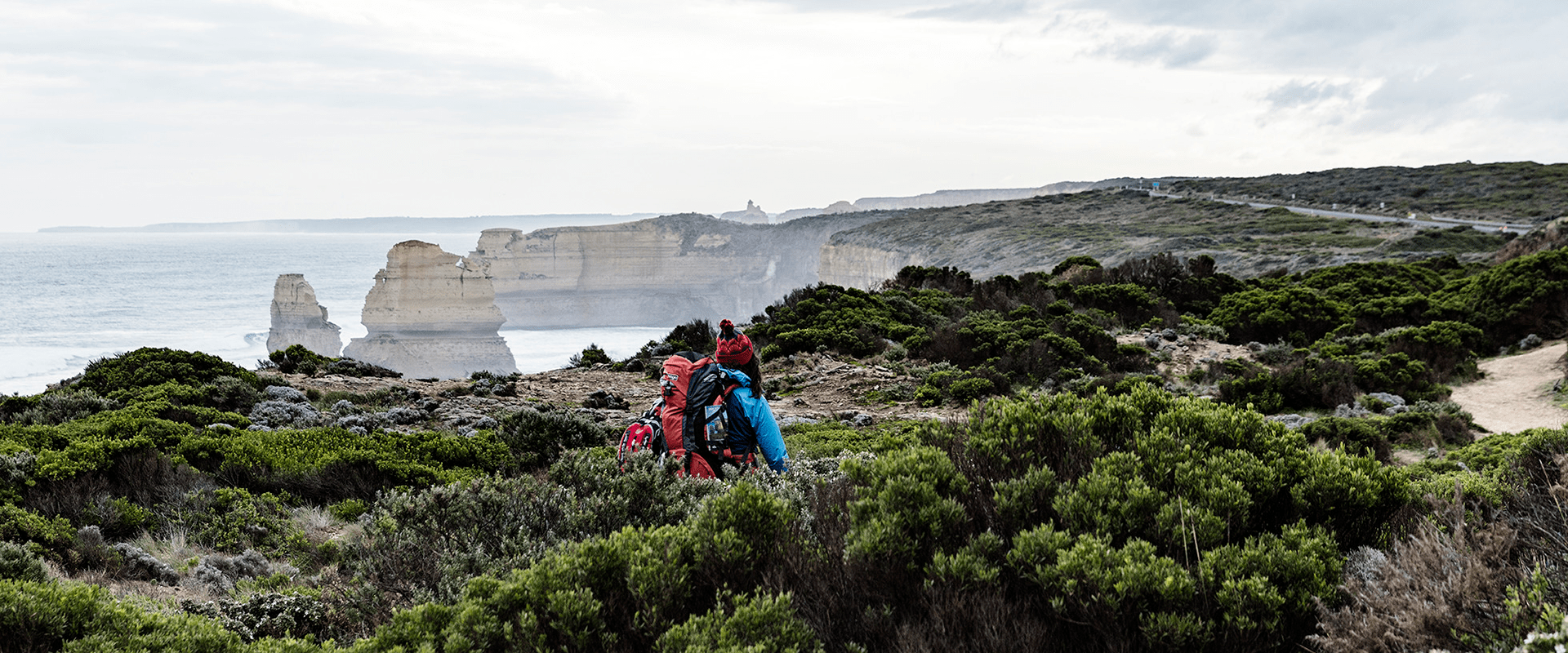 Two hikers walk through coastal shrubbery along a hiking trail with the Twelve Apostles appearing in the background with ocean mist making it's way up steep ocean cliffs.
