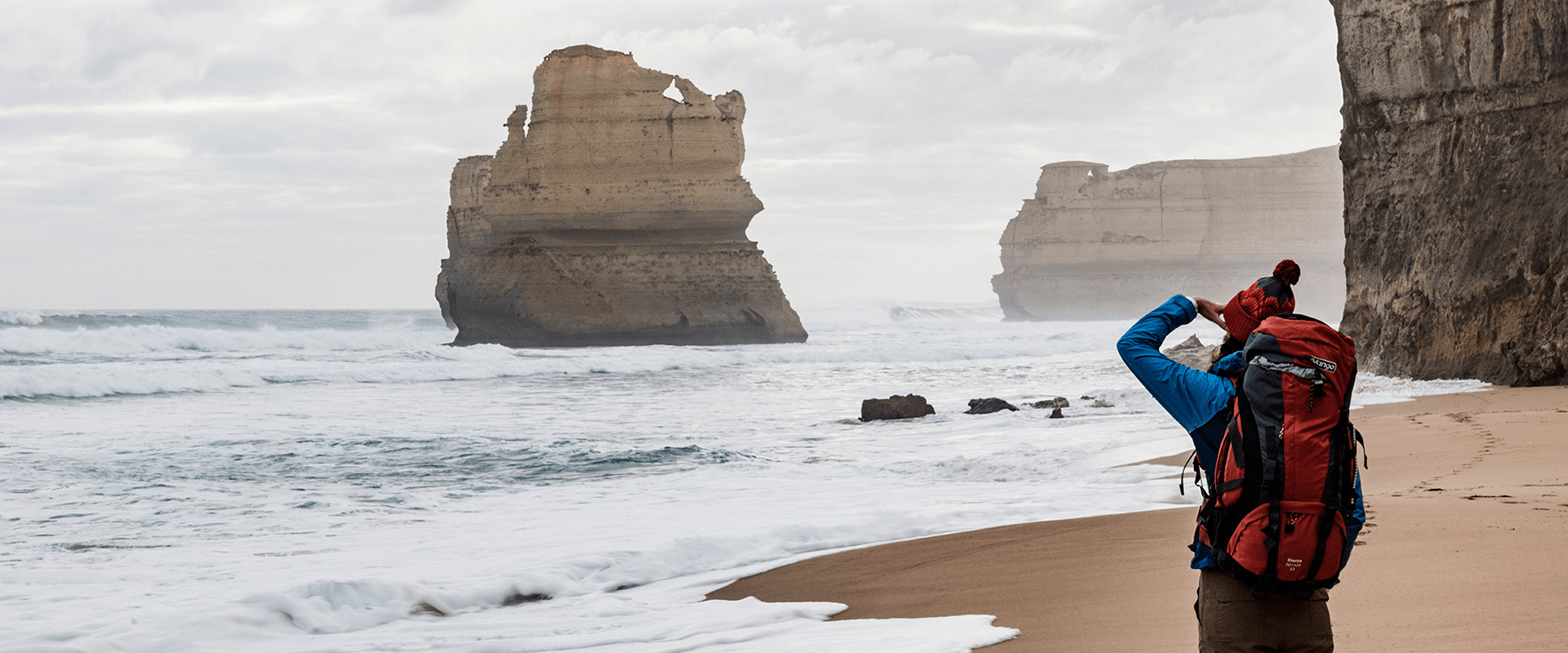 A hiker wears a beanie and a red hiking backpack looks out at the Twelve Apostles to an ocean vista on a sandy beach.